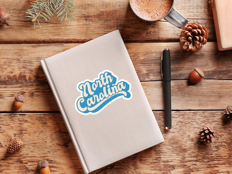 May include: A white sticker with blue text that says "North Carolina" in a retro font style. The sticker is on a white notebook.