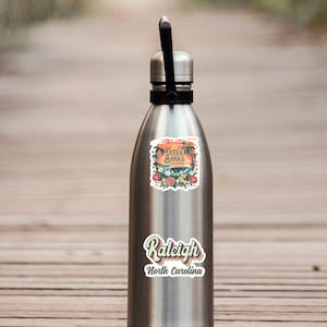 May include: A silver water bottle with two stickers. The top sticker says "Outer Banks" with a sunset and palm trees. The bottom sticker says "Raleigh, North Carolina" in a retro font.