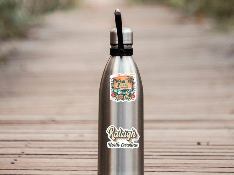 May include: A silver water bottle with two stickers. The top sticker says "Outer Banks" with a sunset and palm trees. The bottom sticker says "Raleigh, North Carolina" in a retro font.