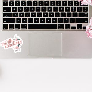 May include: A laptop computer with a sticker of the state of North Carolina with the words "north carolina" in pink lettering and pink floral accents.