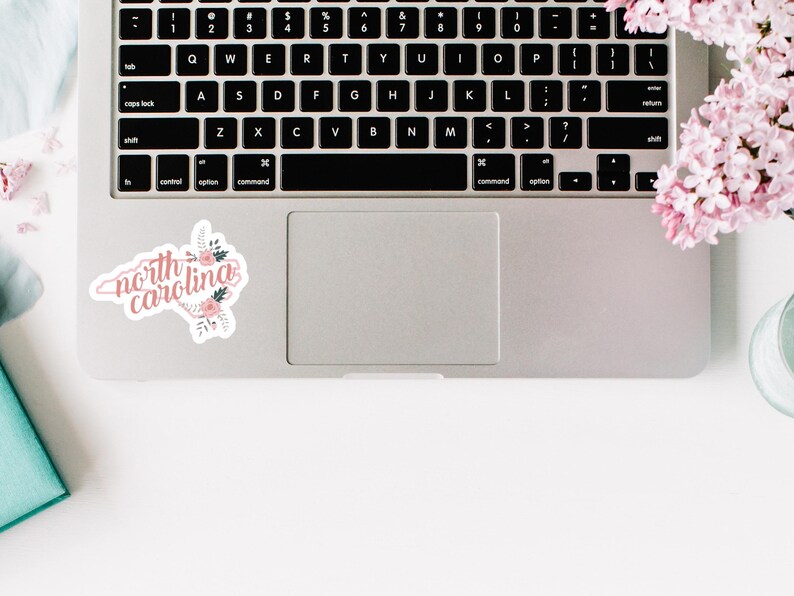 May include: A laptop computer with a sticker of the state of North Carolina with the words "north carolina" in pink lettering and pink floral accents.