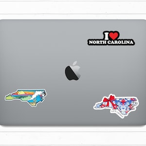 May include: A laptop computer with three North Carolina themed stickers. One sticker is a colorful outline of the state with a sunset and ocean scene. Another sticker is a red, white, and blue patterned outline of the state with a red bow. The third sticker says "I love North Carolina" with a red heart.