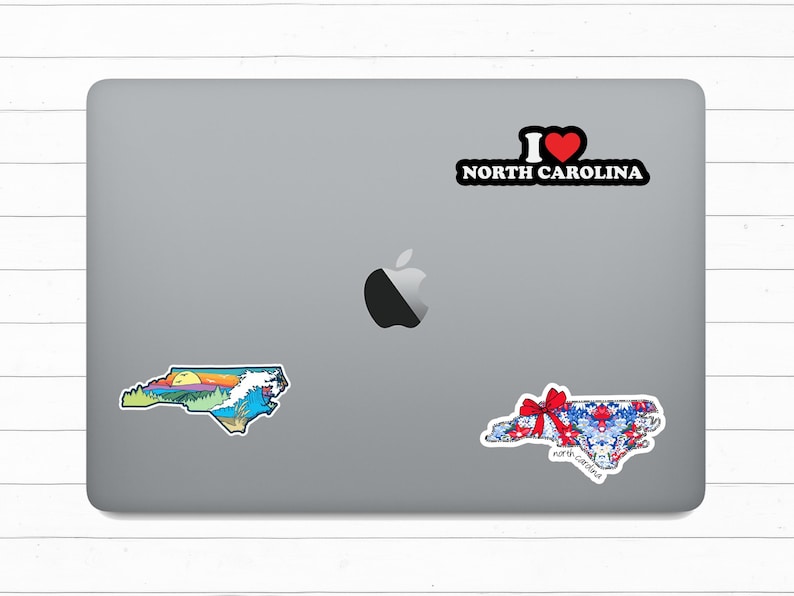 May include: A laptop computer with three North Carolina themed stickers. One sticker is a colorful outline of the state with a sunset and ocean scene. Another sticker is a red, white, and blue patterned outline of the state with a red bow. The third sticker says "I love North Carolina" with a red heart.