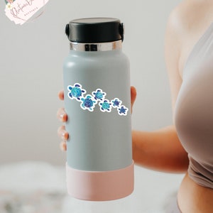 Puede incluir: Una botella de agua reutilizable gris con fondo rosa y una pegatina de un grupo de tortugas marinas azules y verdes nadando en l&iacute;nea.