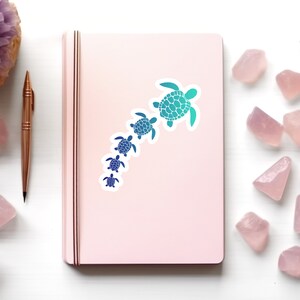 May include: A pink notebook with a sticker of a line of blue and green sea turtles of different sizes. The turtles are facing the same direction and are arranged in a line from largest to smallest.