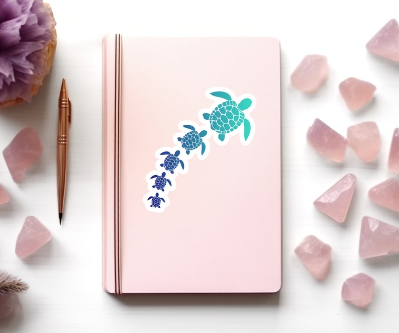 May include: A pink notebook with a sticker of a line of blue and green sea turtles of different sizes. The turtles are facing the same direction and are arranged in a line from largest to smallest.