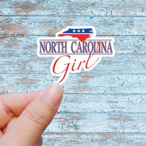 May include: A white sticker with a blue and red outline of the state of North Carolina with stars on the blue portion. The text "NORTH CAROLINA Girl" is written in red and blue letters.