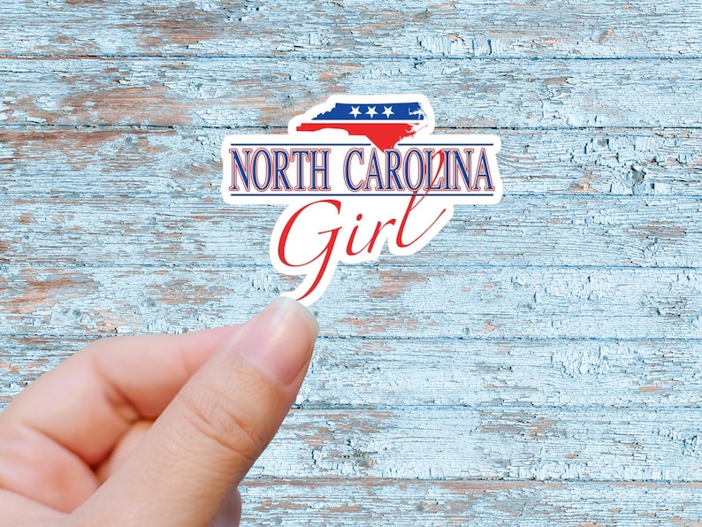 May include: A white sticker with a blue and red outline of the state of North Carolina with stars on the blue portion. The text "NORTH CAROLINA Girl" is written in red and blue letters.