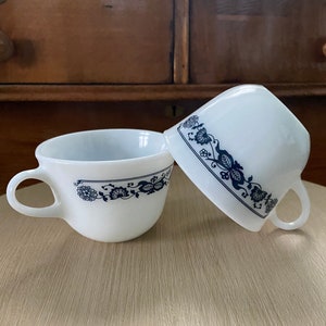 May include: Two white ceramic teacups with blue floral designs. The cups are stacked on top of each other.