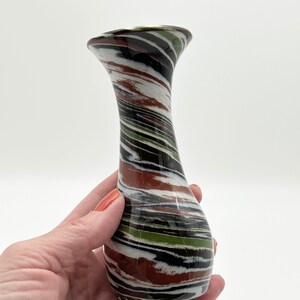 May include: A decorative vase with a unique, swirled pattern of colors including brown, white, black, and green. The vase has a flared top and a rounded base. The vase is held in a hand against a white background.