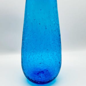 May include: A tall, blue glass vase with a textured, crackled surface. The vase is a light blue color and has a rounded bottom.