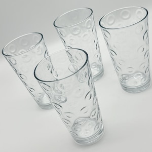May include: Four clear, tall drinking glasses with a textured pattern of circles and curved lines. The glasses are empty and arranged at an angle against a white background. The glasses are made of clear glass.