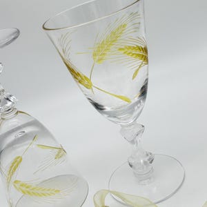 May include: Two clear wine glasses with a wheat stalk design in yellow. The glasses have a clear stem and base. A gold beaded ribbon is in the foreground.