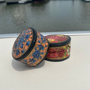 May include: Two small round decorative boxes with intricate floral designs. The boxes are painted in black, gold, blue, red, and pink. The boxes are made of wood and have a glossy finish.