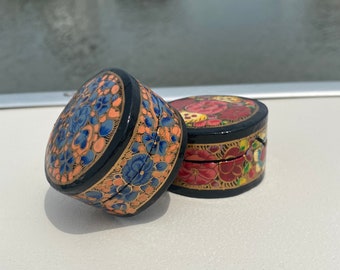May include: Two small round decorative boxes with intricate floral designs. The boxes are painted in black, gold, blue, red, and pink. The boxes are made of wood and have a glossy finish.