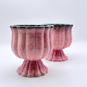 May include: Two ceramic pedestal bowls featuring a fluted, scalloped design. The exterior has a mottled pink glaze with a darker, speckled finish near the rim and base. The interior rim is a deep, dark green, contrasting with the pink body.