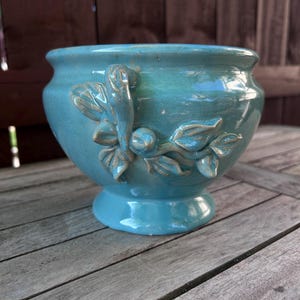 May include: A turquoise ceramic planter with a pedestal base and a glossy finish. The planter features a raised floral design on the side. The planter is designed for indoor or outdoor use.
