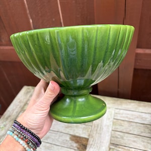 May include: A glossy, forest green ceramic bowl with a pedestal base and vertical ribbed detailing along the exterior. The bowl features a wide, flared rim and a smooth, reflective finish that catches the light, creating subtle variations in the green tone.