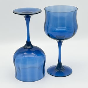 May include: Two cobalt blue wine glasses. One glass is upright, the other is inverted. The glasses have a tulip-shaped bowl and a long stem. The base of each glass is round. The background is white.
