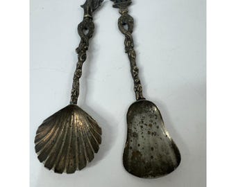 Könnte beinhalten: Zwei silberfarbene Metalllöffel mit angelaufenen Oberflächen. Der linke Löffel hat eine muschelförmige Laffe, der rechte eine birnenförmige. Beide Griffe sind mit kunstvollen, gedrehten Metallverzierungen und kleinen Figuren versehen.