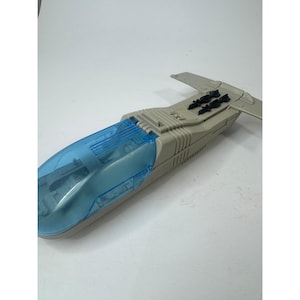 Pode incluir: Uma nave espacial de brinquedo em plástico cinzento com uma cobertura de cockpit azul translúcida e dois pequenos acessórios de mísseis pretos montados na parte traseira da fuselagem. O veículo tem um corpo aerodinâmico e alongado.