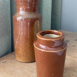 以下が含まれることがあります： 2つの茶色の陶器の壺。1つは他方より背が高く、表面はざらざらとした質感です。壺は木製の表面の上に置かれています。