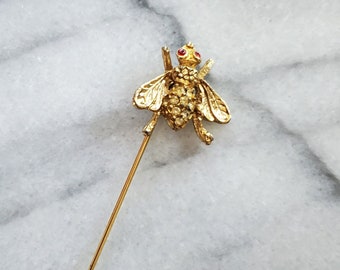May include: A gold-colored bee pin with a long, slender needle. The bee's body is encrusted with small, clear crystals, and its eyes are red. The pin is set against a white and gray marble background.
