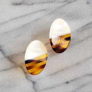 May include: A pair of oval-shaped stud earrings. Each earring is divided into two sections: a white top and a bottom section with a striped pattern in shades of brown and amber. The earrings are set against a marble surface.