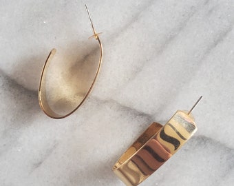 May include: Two gold-colored earrings. One is a classic oval hoop, and the other is a rectangular hoop. The earrings are displayed on a white and gray marble surface. The earrings have a shiny, metallic finish.