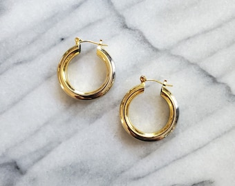 May include: A pair of gold-colored hoop earrings. The earrings are thick and have a rounded shape. They are displayed on a white and gray marble surface.