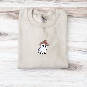 May include: A beige sweatshirt with a crew neck. The sweatshirt features an embroidered design of a white ghost wearing a brown cowboy hat. The ghost has black eyes and a smiling mouth. The sweatshirt is folded on a light wood surface.