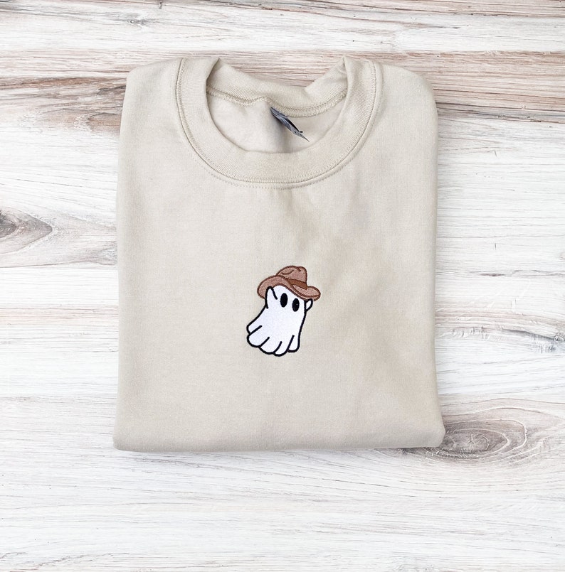 May include: A beige sweatshirt with a crew neck. The sweatshirt features an embroidered design of a white ghost wearing a brown cowboy hat. The ghost has black eyes and a smiling mouth. The sweatshirt is folded on a light wood surface.