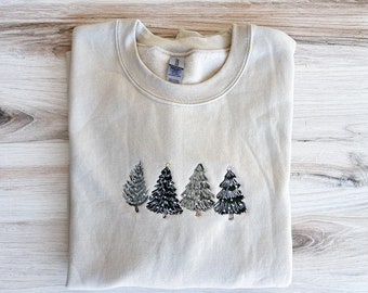 Puede incluir: Sudadera beige con un diseño bordado de cuatro árboles de Navidad en tonos verdes, grises y negros. Los árboles están dispuestos en una fila en el pecho. La sudadera está doblada sobre una superficie de madera clara.
