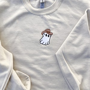 May include: A beige sweatshirt with a crew neck and long sleeves. The sweatshirt features an embroidered patch of a white ghost wearing a brown cowboy hat. The ghost has black eyes and a smiling mouth.