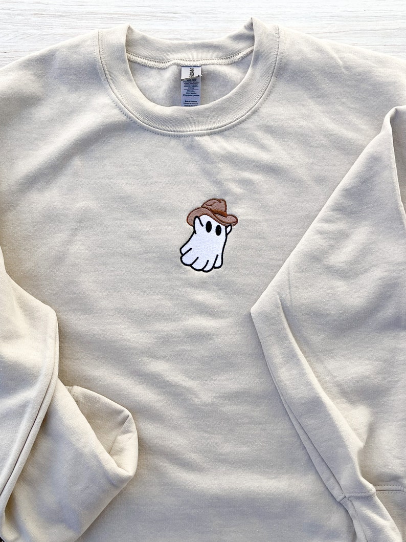 May include: A beige sweatshirt with a crew neck and long sleeves. The sweatshirt features an embroidered patch of a white ghost wearing a brown cowboy hat. The ghost has black eyes and a smiling mouth.
