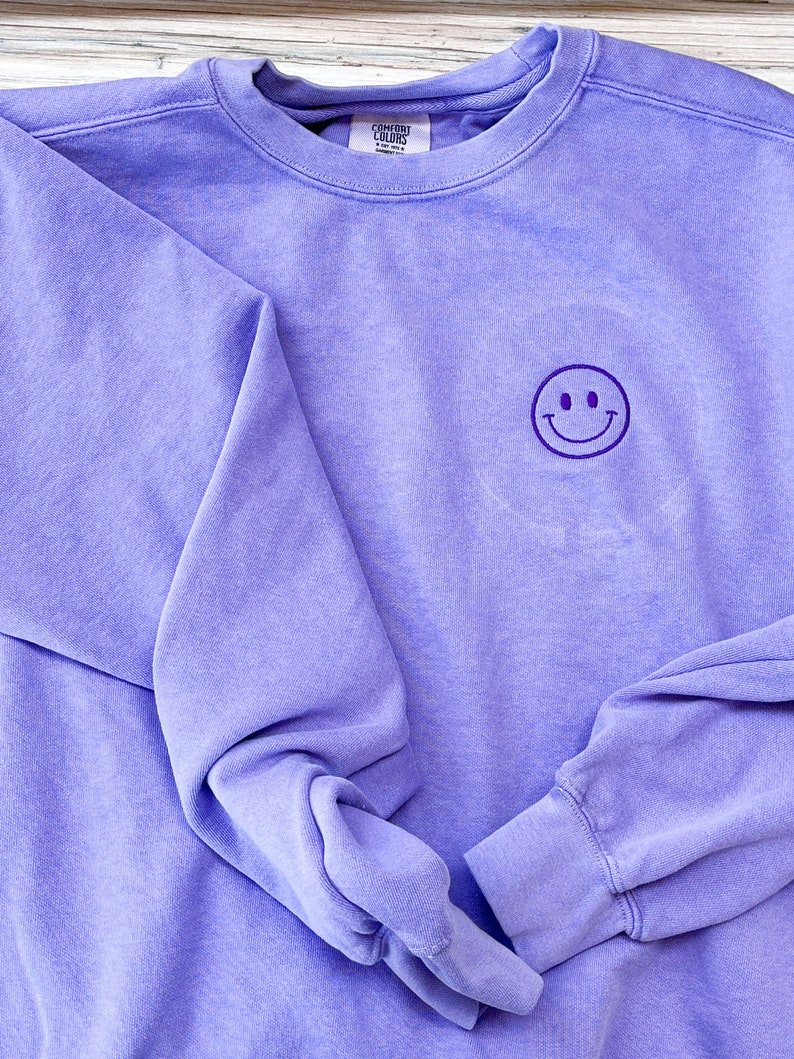 May include: A lavender sweatshirt with a purple embroidered smiley face on the front. The sweatshirt is folded in half and the tag reads "Comfort Colors".