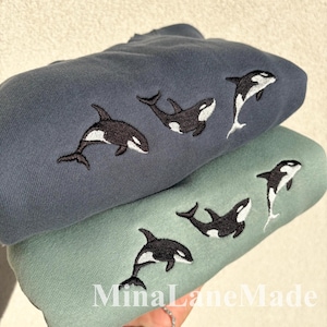 May include: Two sweatshirts, one blue-grey and one sage green, are stacked. Each sweatshirt has embroidered orca whale designs in black and white. The text "MinaLaneMade" is visible at the bottom.