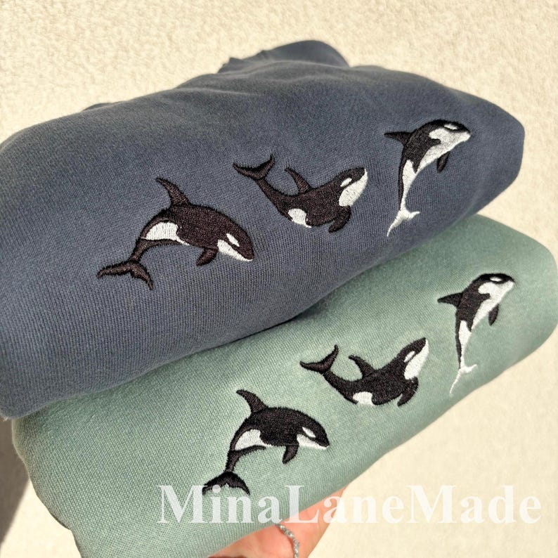 May include: Two sweatshirts, one blue-grey and one sage green, are stacked. Each sweatshirt has embroidered orca whale designs in black and white. The text "MinaLaneMade" is visible at the bottom.