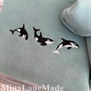 May include: A pale green sweatshirt featuring three embroidered orca whales in black and white. The whales are positioned across the front. The brand name "MinaLaneMade" is printed at the bottom of the image.