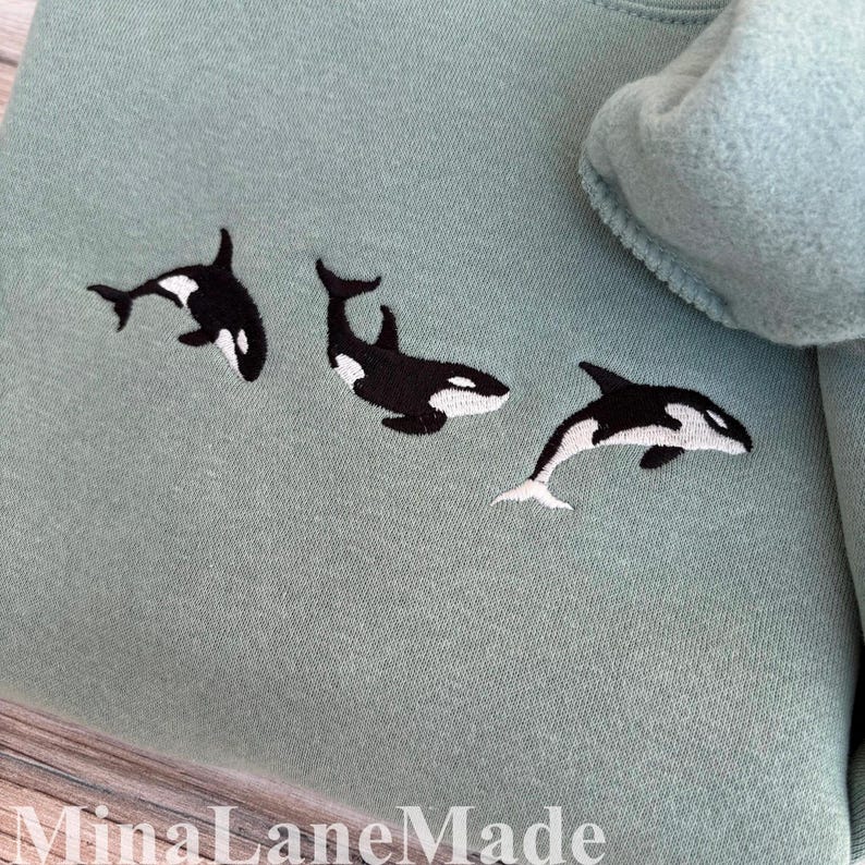 May include: A pale green sweatshirt featuring three embroidered orca whales in black and white. The whales are positioned across the front. The brand name "MinaLaneMade" is printed at the bottom of the image.