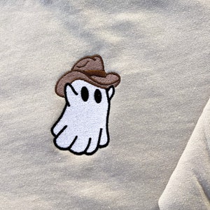 May include: A light beige sweatshirt featuring an embroidered patch of a white ghost wearing a brown cowboy hat. The ghost has black eyes and a black outline. The sweatshirt has a crew neck and long sleeves.