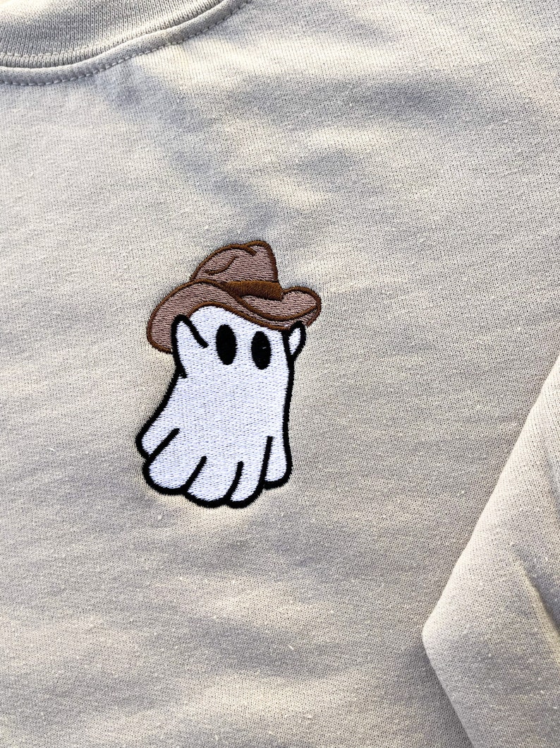 May include: A light beige sweatshirt featuring an embroidered patch of a white ghost wearing a brown cowboy hat. The ghost has black eyes and a black outline. The sweatshirt has a crew neck and long sleeves.