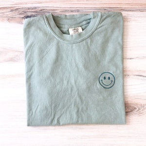 May include: A sage green t-shirt with a blue embroidered smiley face. The t-shirt is made by the brand "Comfort Colors".