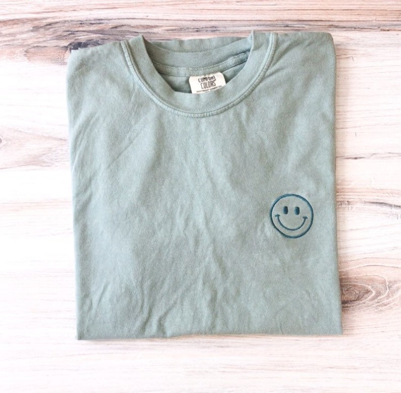 May include: A sage green t-shirt with a blue embroidered smiley face. The t-shirt is made by the brand "Comfort Colors".