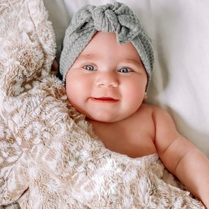 May include: A baby wearing a gray knitted turban with a knot on top, wrapped in a beige fuzzy blanket. The baby is smiling and looking at the camera.