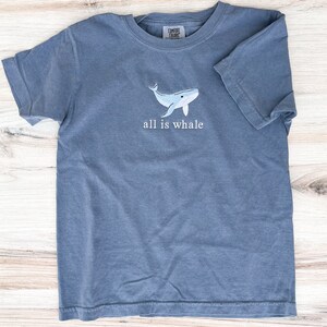 Może przedstawiać: Niebieski t-shirt z kr&oacute;tkim rękawem z haftowanym jasnoniebieskim wzorem wieloryba i napisem "all is whale". Koszulka wykonana jest z miękkiego materiału i ma okrągły dekolt.