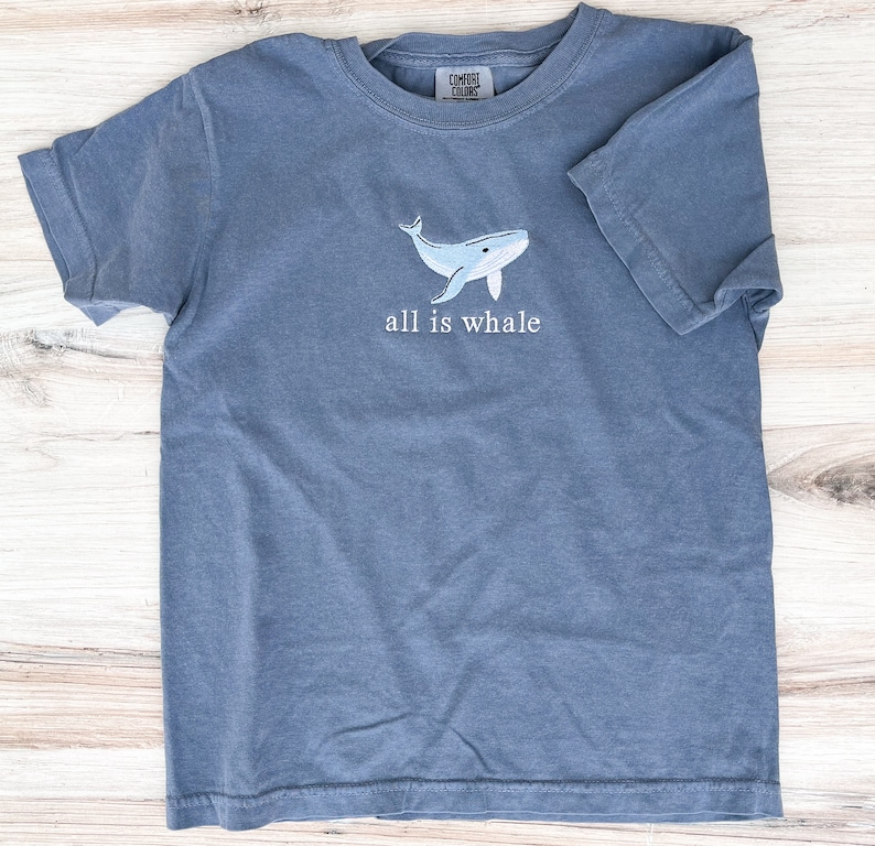 Może przedstawiać: Niebieski t-shirt z kr&oacute;tkim rękawem z haftowanym jasnoniebieskim wzorem wieloryba i napisem "all is whale". Koszulka wykonana jest z miękkiego materiału i ma okrągły dekolt.