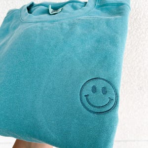 May include: A teal blue sweatshirt with a black embroidered smiley face on the front.