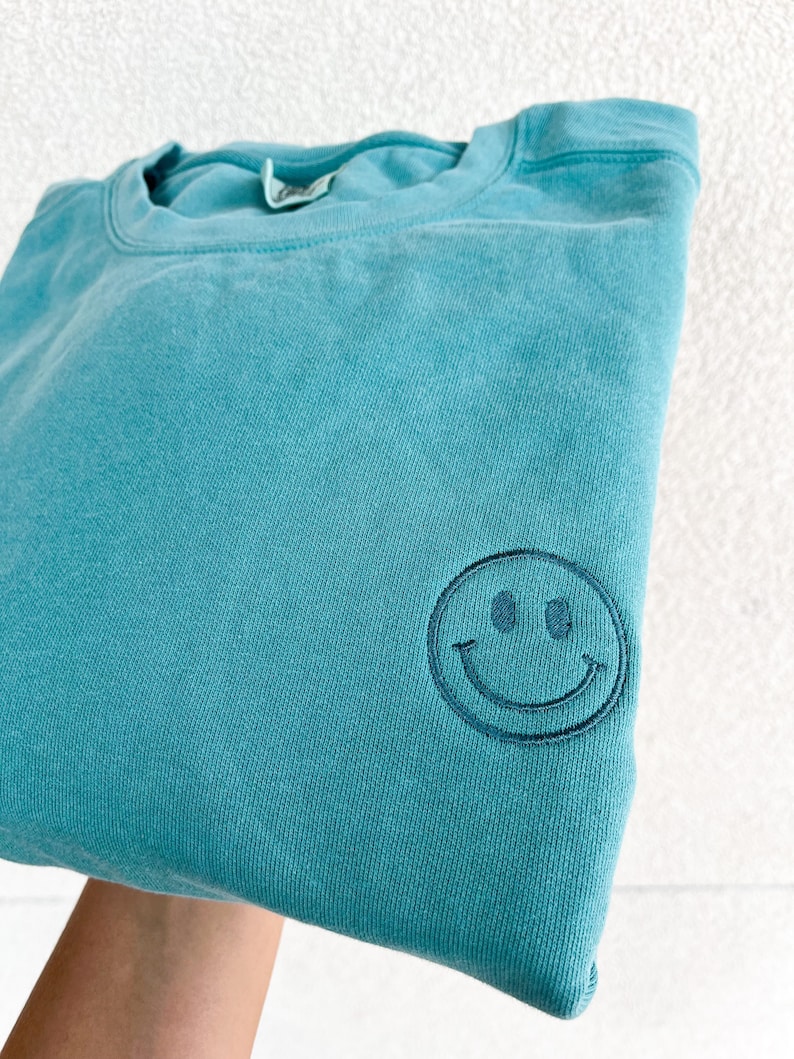 May include: A teal blue sweatshirt with a black embroidered smiley face on the front.