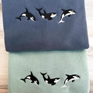 May include: Two sweatshirts, one blue and one green, each embroidered with three black and white orca whale designs. The whales are in different poses, with some leaping and others swimming. The sweatshirts are folded and stacked.